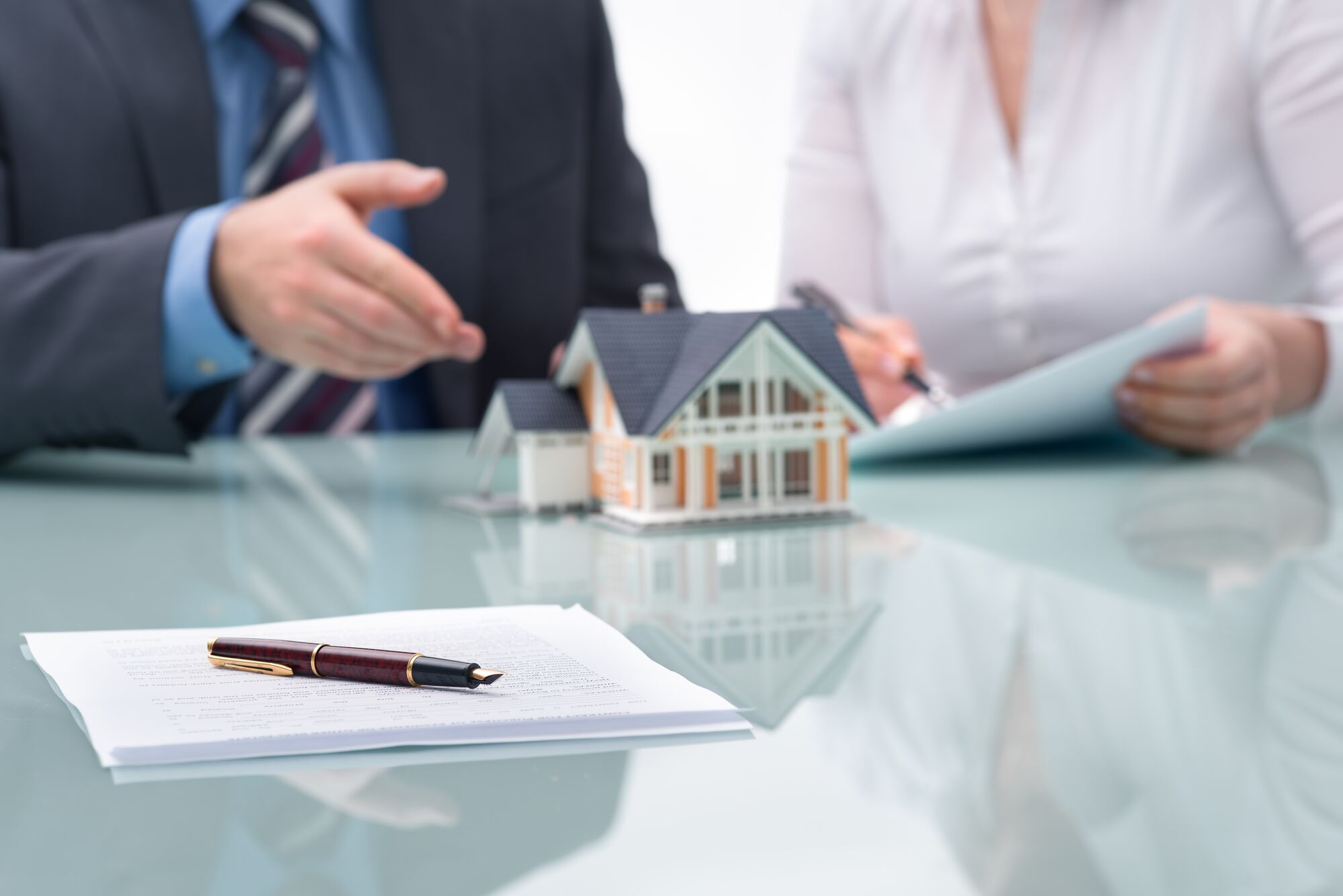 A business professional gestures while discussing real estate, with a model house and documents on the table, highlighting a property transaction.